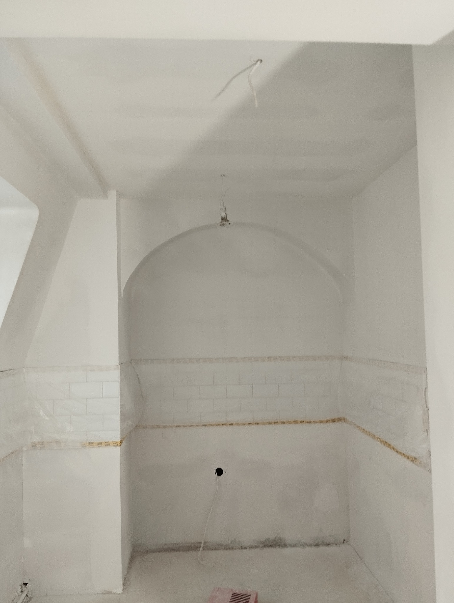 Biała łazienka w trakcie remontu: częściowo ułożone białe płytki, ściany przygotowane do malowania, widoczne kable oświetleniowe. Łuk nad fragmentem ściany z płytkami.