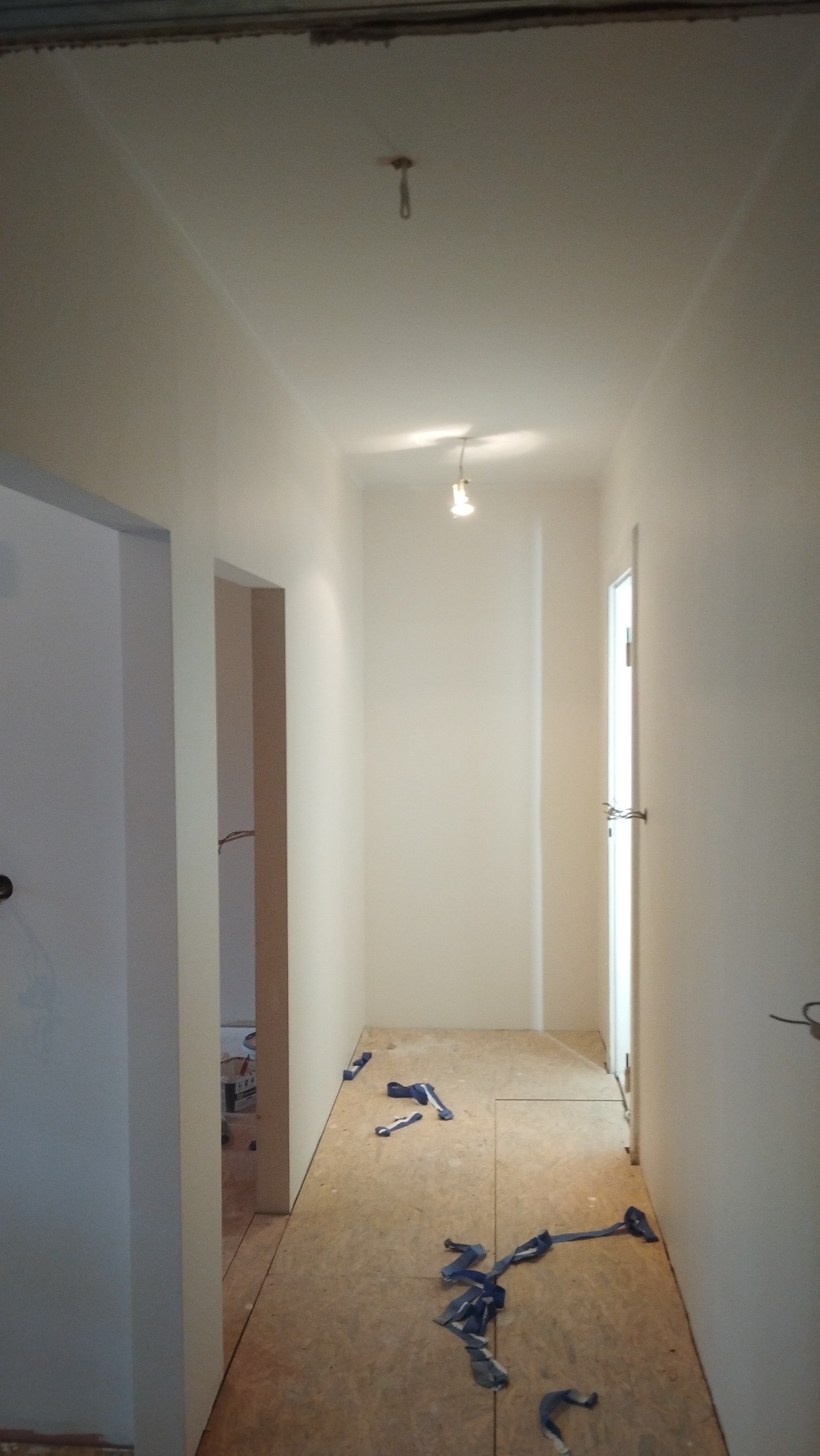 Jasny korytarz po malowaniu ścian na biało. Na podłodze płyty OSB i resztki taśmy malarskiej. Widoczne drzwi i fragmenty innych pomieszczeń. Oświetlenie sufitowe.
