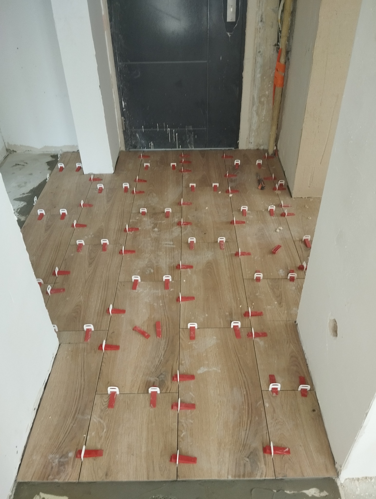 Układanie drewnopodobnych płytek podłogowych z użyciem czerwonych klipsów poziomujących, widok z góry na świeżo ułożoną podłogę w korytarzu, ciemne drzwi w tle.