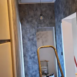Wnętrze po odświeżeniu, ściany z szarą tapetą o geometrycznym wzorze, metalowa drabina na pierwszym planie, widoczna lampa wisząca i fragment otwartego przejścia do innego pomieszczenia, podłoga...