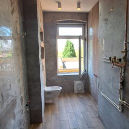 Andrzej Makowiak - Wykończona łazienka z szarymi płytkami imitującymi marmur na ścianach i drewnopodobnymi płytkami na podłodze. Wisząca toaleta i okno z widokiem na zieleń. Widoczne rury miedziane.