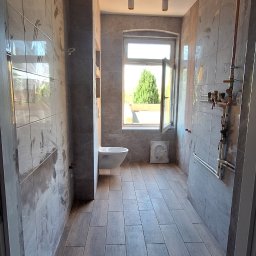 Andrzej Makowiak - Wykończona łazienka z szarymi płytkami imitującymi drewno na podłodze, szare płytki na ścianach, podwieszana toaleta, okno z widokiem na zieleń i nowoczesne oświetlenie punktowe na suficie.