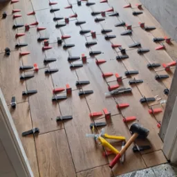 Układanie drewnopodobnych płytek podłogowych w łazience z użyciem systemu poziomowania, widoczne narzędzia: młotek, nóż oraz szczypce do klinów.