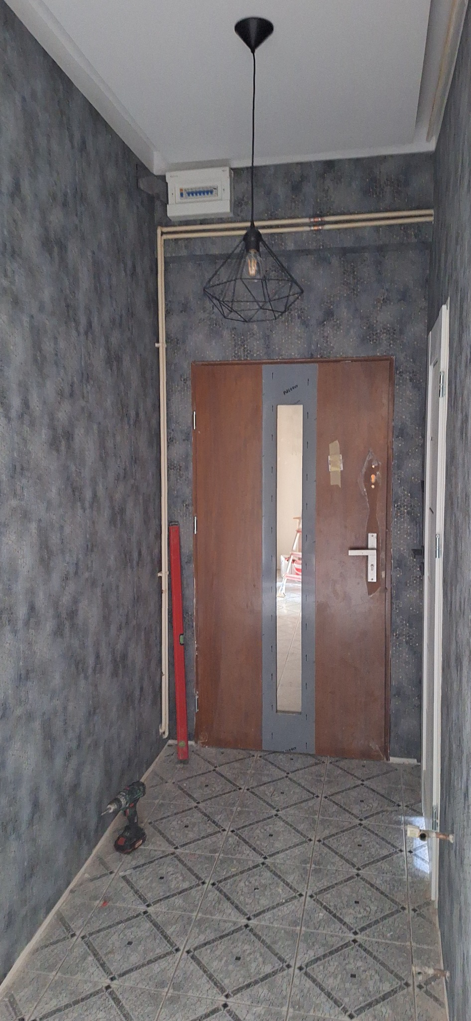 Korytarz w trakcie remontu: brązowe drzwi z szybą zabezpieczone szarą folią, poziomica oparta o ścianę, wiertarka na podłodze z szarych płytek w romby, surowa żarówka w metalowej oprawie zwisa...