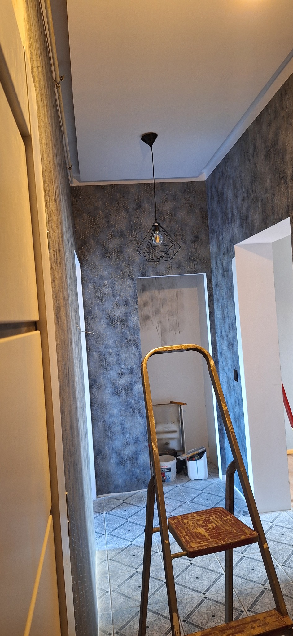 Wnętrze po odświeżeniu, ściany z szarą tapetą o geometrycznym wzorze, metalowa drabina na pierwszym planie, widoczna lampa wisząca i fragment otwartego przejścia do innego pomieszczenia, podłoga...