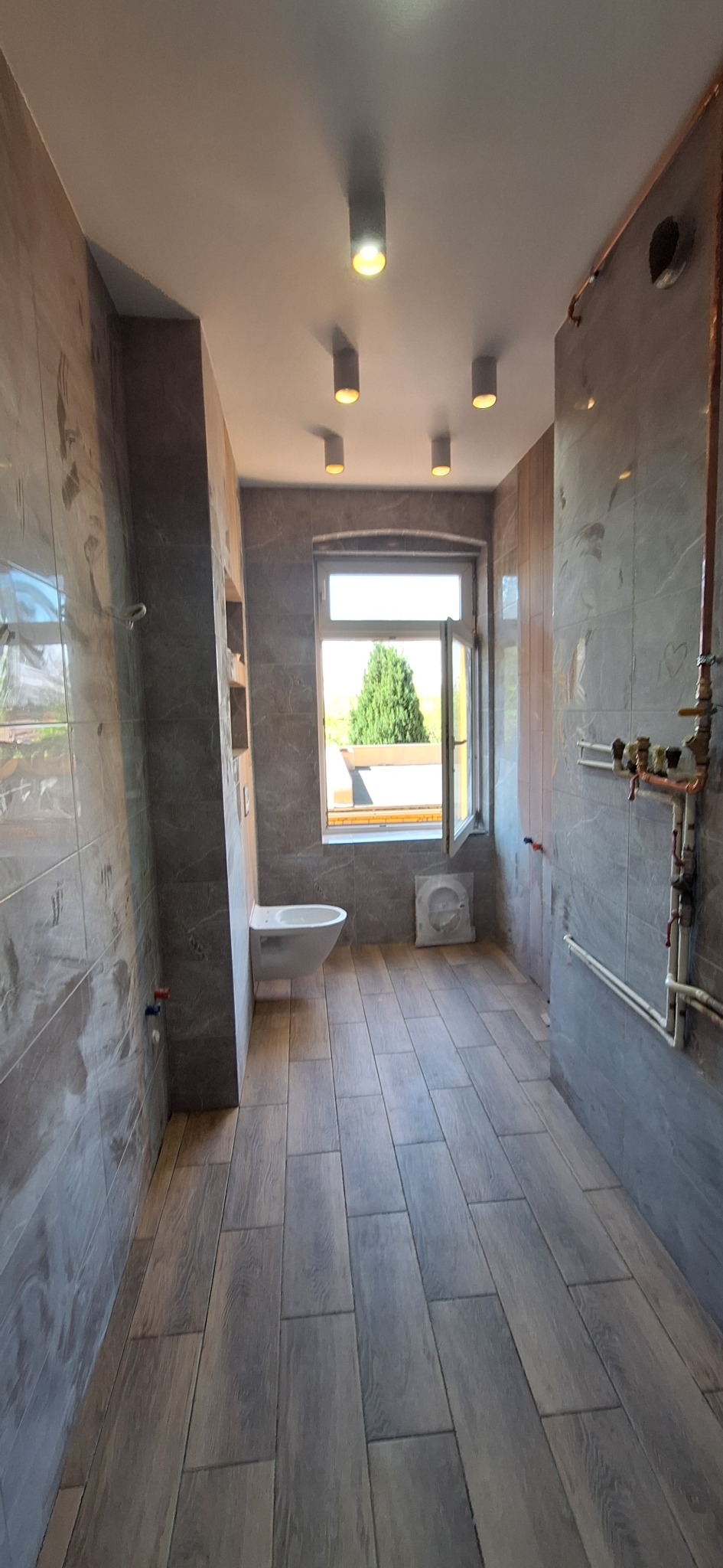 Wykończona łazienka z szarymi płytkami imitującymi marmur na ścianach i drewnopodobnymi płytkami na podłodze. Wisząca toaleta i okno z widokiem na zieleń. Widoczne rury miedziane.
