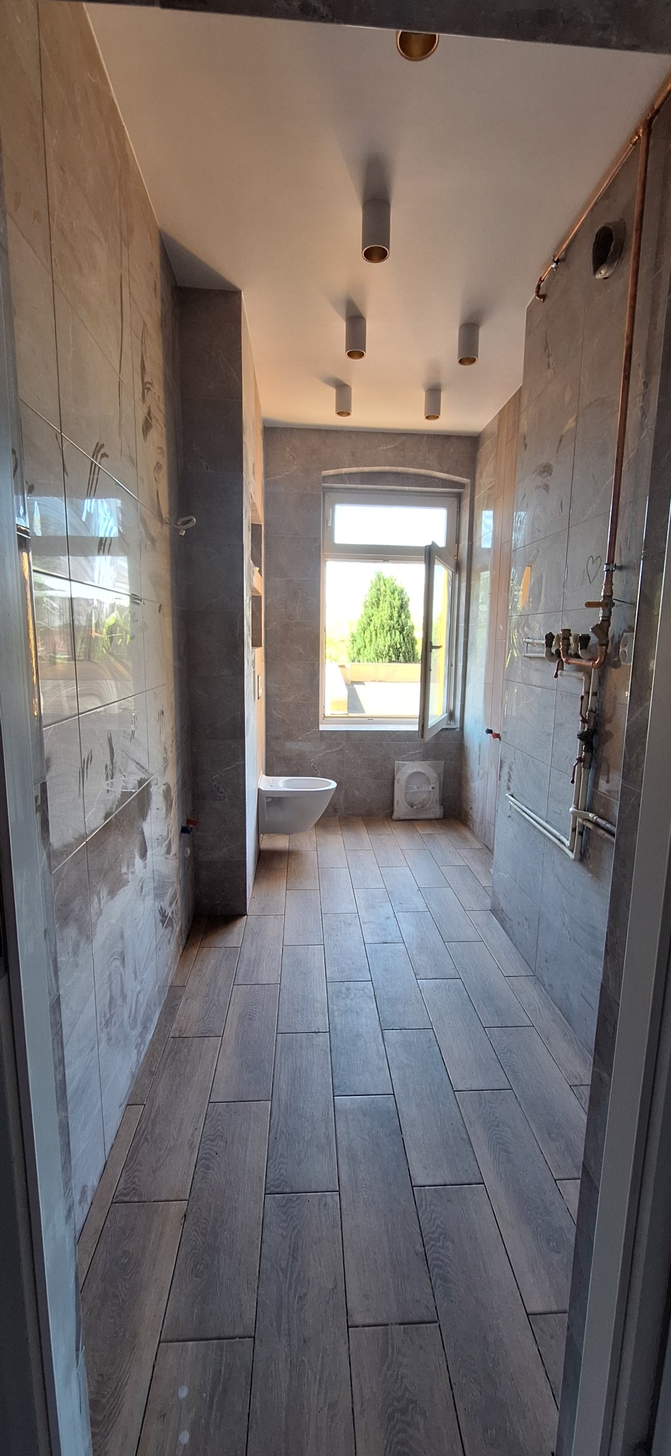 Wykończona łazienka z szarymi płytkami imitującymi drewno na podłodze, szare płytki na ścianach, podwieszana toaleta, okno z widokiem na zieleń i nowoczesne oświetlenie punktowe na suficie.