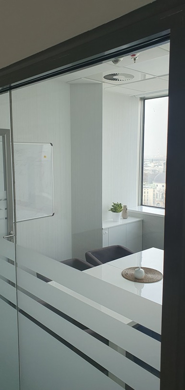 Nowoczesne biuro z przeszkloną ścianą z poziomymi pasami, biały stół z dekoracją, szare krzesła i widok na miasto za oknem.