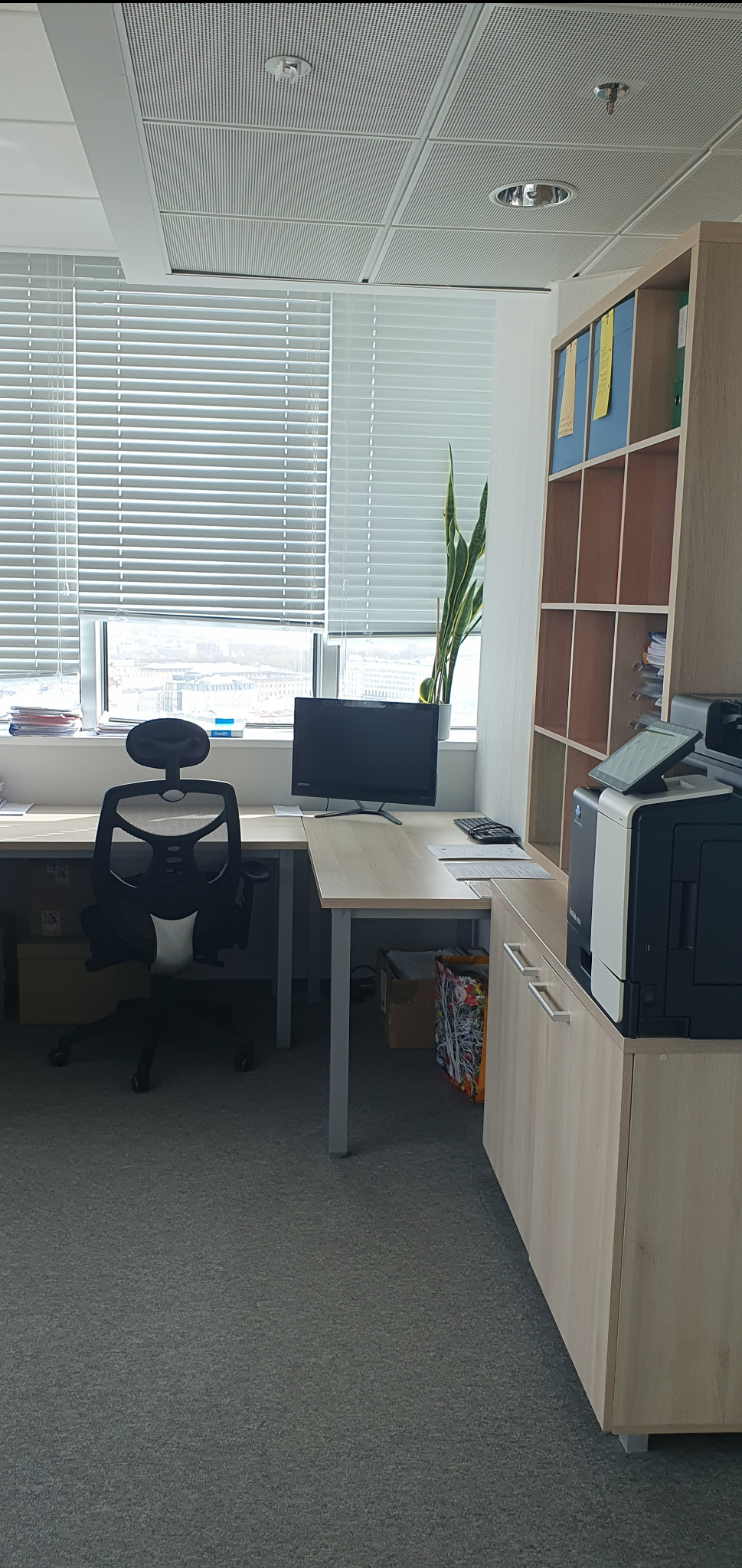 Jasne biuro w Warszawie z biurkiem, krzesłem, komputerem, drukarką i regałem na dokumenty, widok zza okna z żaluzjami.