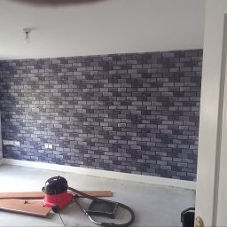 Tab-bud - Pokój z tapetą imitującą cegłę, widok przez uchylone drzwi, na podłodze leżą narzędzia i odkurzacz.