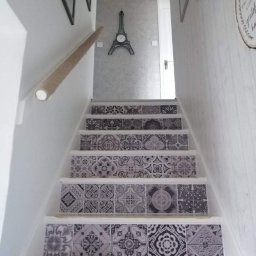Tab-bud - Schody wyłożone dekoracyjnymi płytkami w odcieniach szarości i bieli, z motywami geometrycznymi i kwiatowymi, prowadzące w górę, z widoczną poręczą obłożoną jutą i dekoracją ścienną w kształcie...