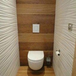 Tab-bud - Nowoczesna toaleta z drewnianą boazerią i trójwymiarowymi białymi panelami ściennymi, podwieszana miska WC, drewniana podłoga, widoczny dozownik papieru toaletowego i szczotka do WC.