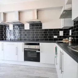 Nowoczesna kuchnia z białymi szafkami i czarnymi płytkami ściennymi ułożonymi w cegiełkę, trzy srebrne okapy nad płytą grzewczą, jasnoszara podłoga.