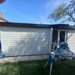 Tab-bud - Nowoczesny, szary domek narzędziowy z czarnym dachem, częściowo wykończony, z otwartymi drzwiami, przez które widać pracownika; na pierwszym planie pilarka stołowa na trawie.