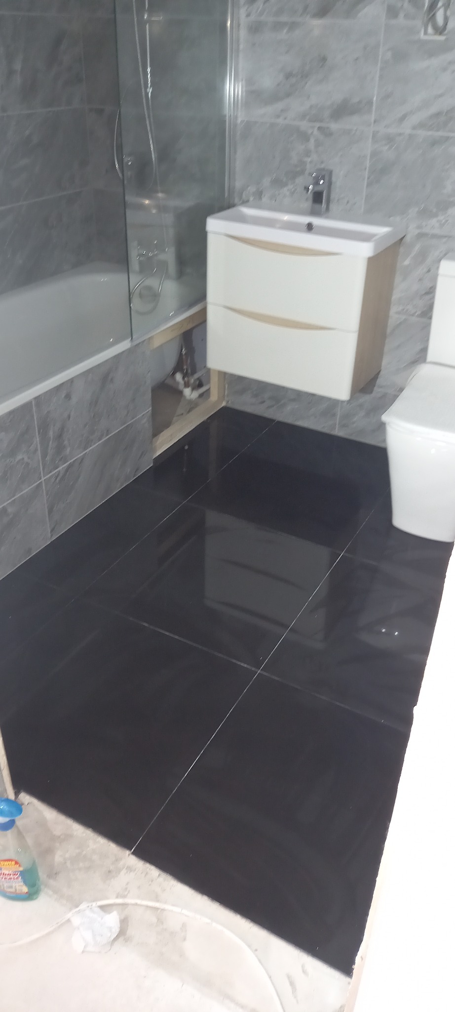 Ciemne płytki podłogowe w łazience z umywalką na szafce, wanną za szklaną ścianką i toaletą, widoczne niedokończone prace wykończeniowe przy podłodze i rurach.