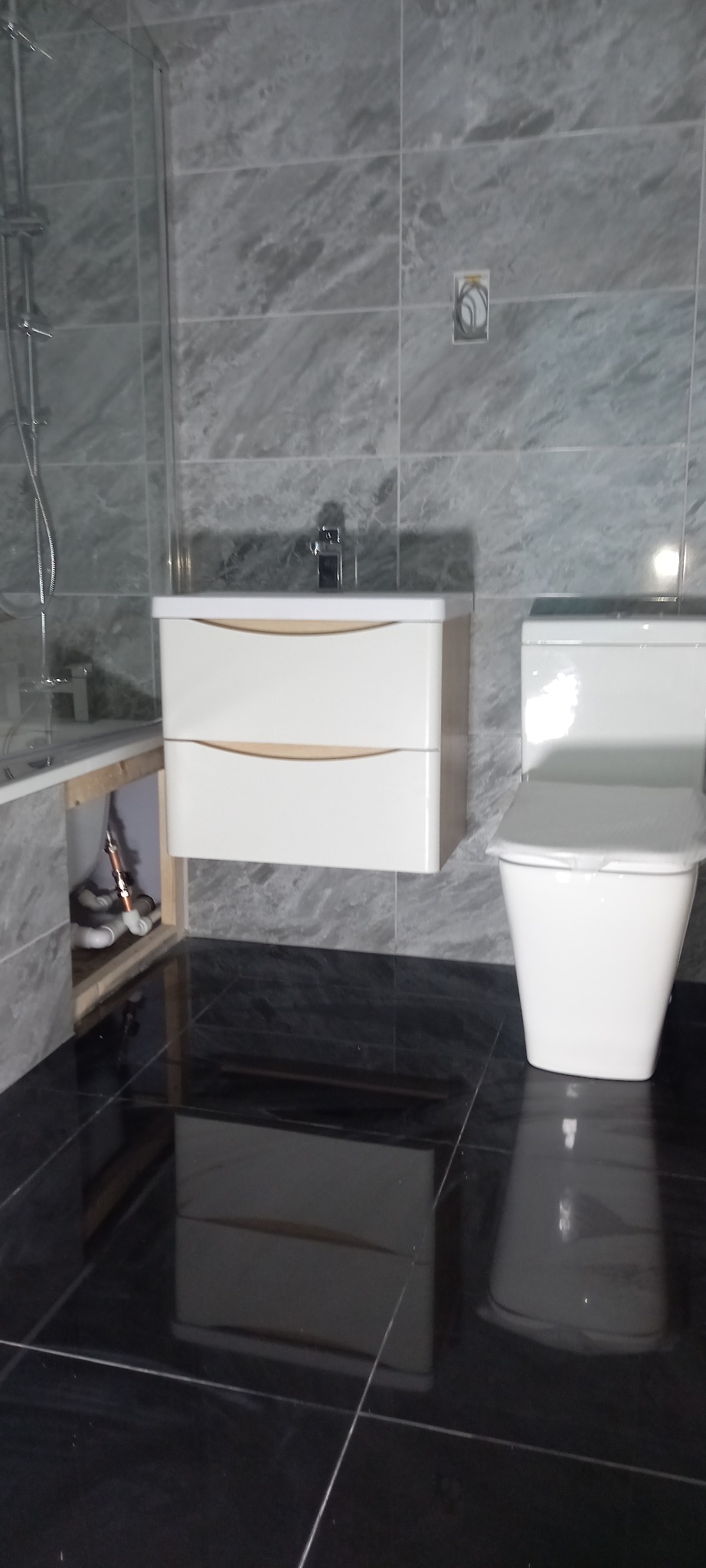 Nowoczesna łazienka z marmurowymi płytkami, białą szafką pod umywalką i czarną, błyszczącą podłogą; widoczne rury pod wanną.