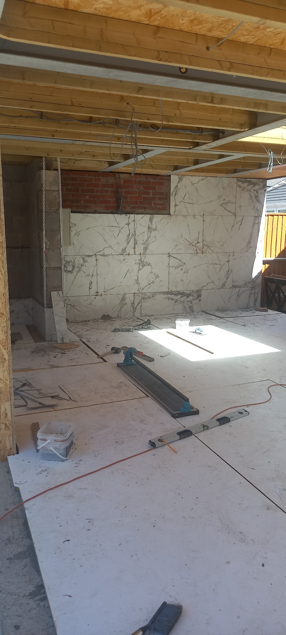Wnętrze w trakcie układania płytek imitujących marmur na ścianie, widoczny niezabudowany sufit z drewnianych belek, narzędzia glazurnicze na podłodze, częściowo wykończona ściana z cegły.