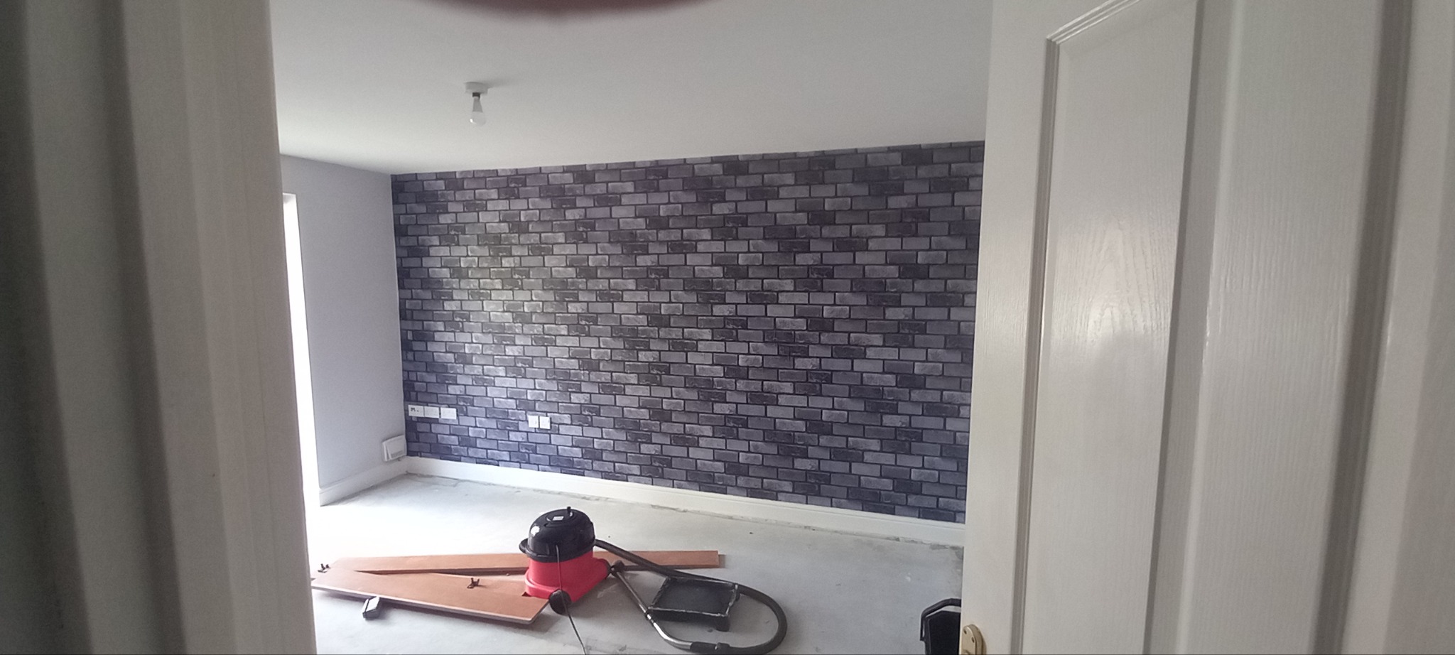Pokój z tapetą imitującą cegłę, widok przez uchylone drzwi, na podłodze leżą narzędzia i odkurzacz.
