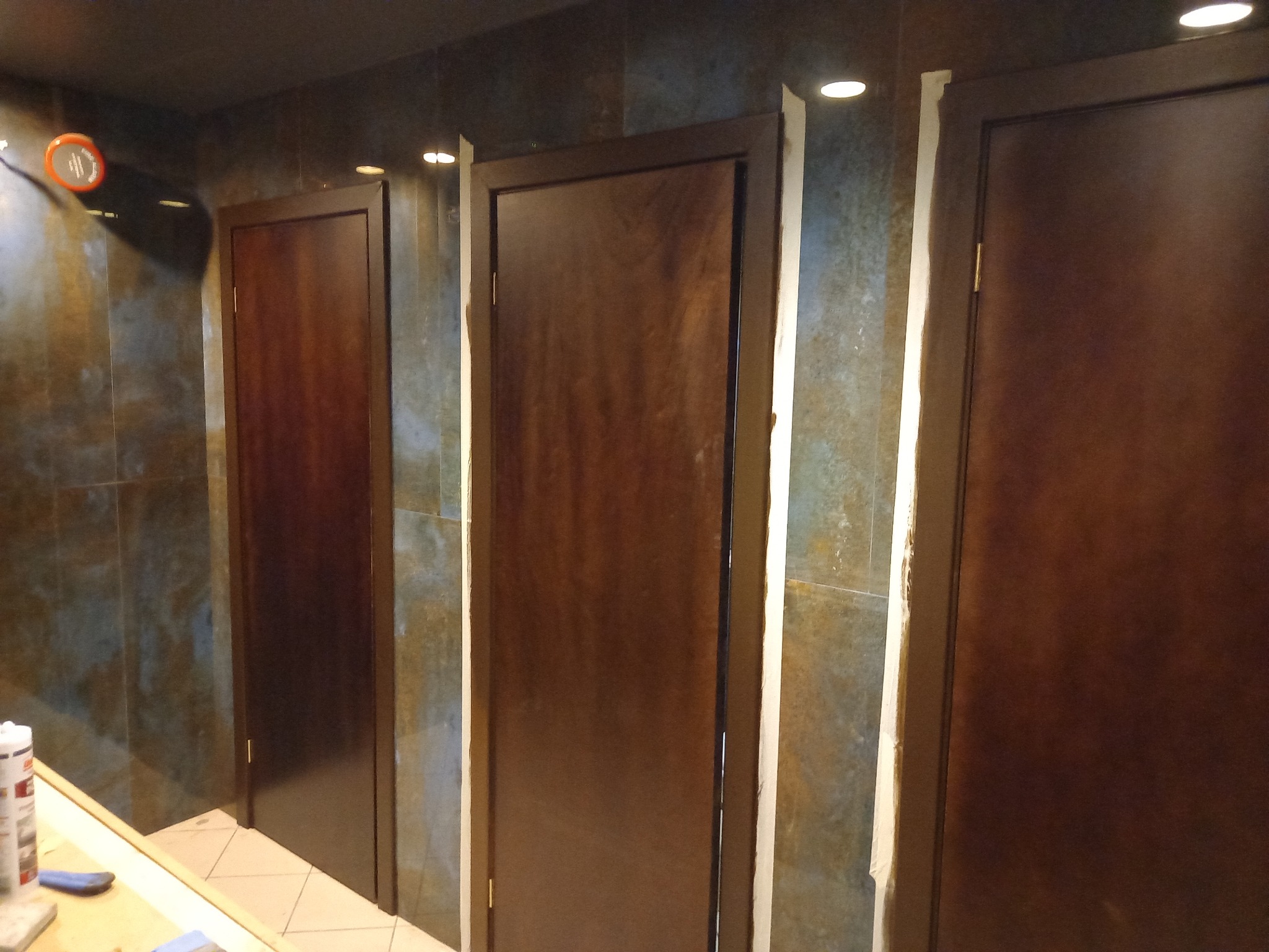 Trzy ciemnobrązowe drzwi z ościeżnicami w trakcie montażu lub obróbki wykończeniowej, widoczne ślady gipsu lub szpachli, ściany wyłożone płytkami imitującymi kamień w odcieniach brązu i szarości...