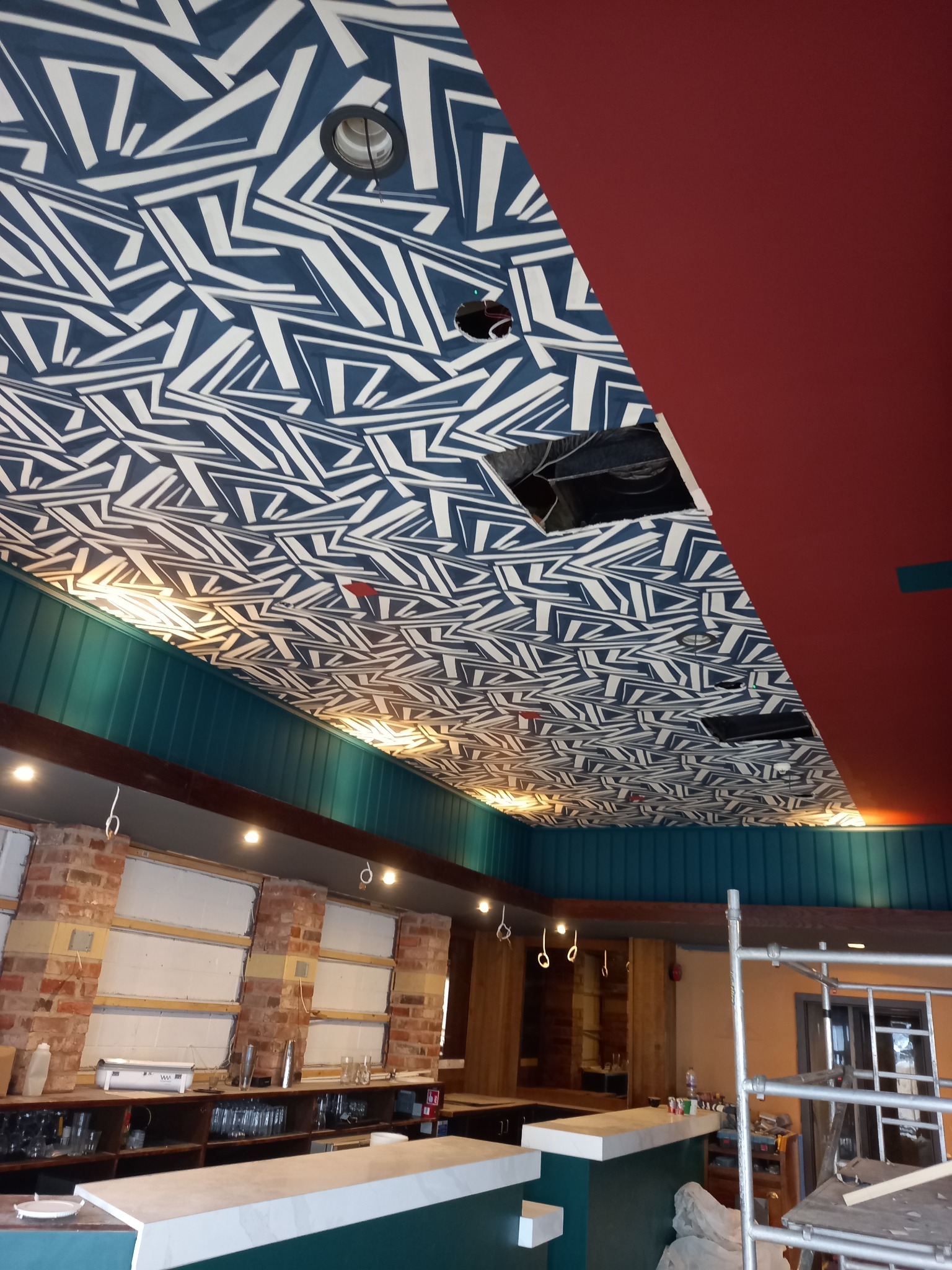 Nietypowy sufit w lokalu z geometrycznym wzorem w odcieniach bieli i granatu, widoczne otwory instalacyjne i fragmenty ścian w kolorze bordowym i morskim, bar w trakcie remontu.