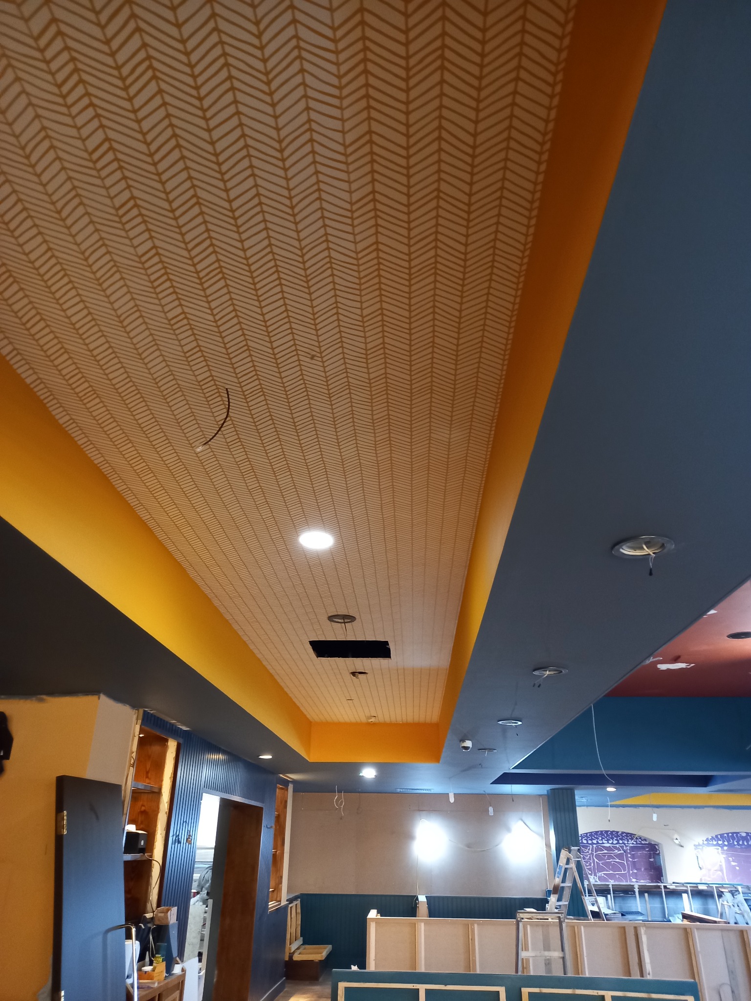 Wnętrze lokalu w trakcie wykańczania: pomalowany sufit z wzorem jodełki, żółte pasy dekoracyjne, częściowo pomalowane ściany na niebiesko i żółto, widoczne instalacje elektryczne i schody malarskie.