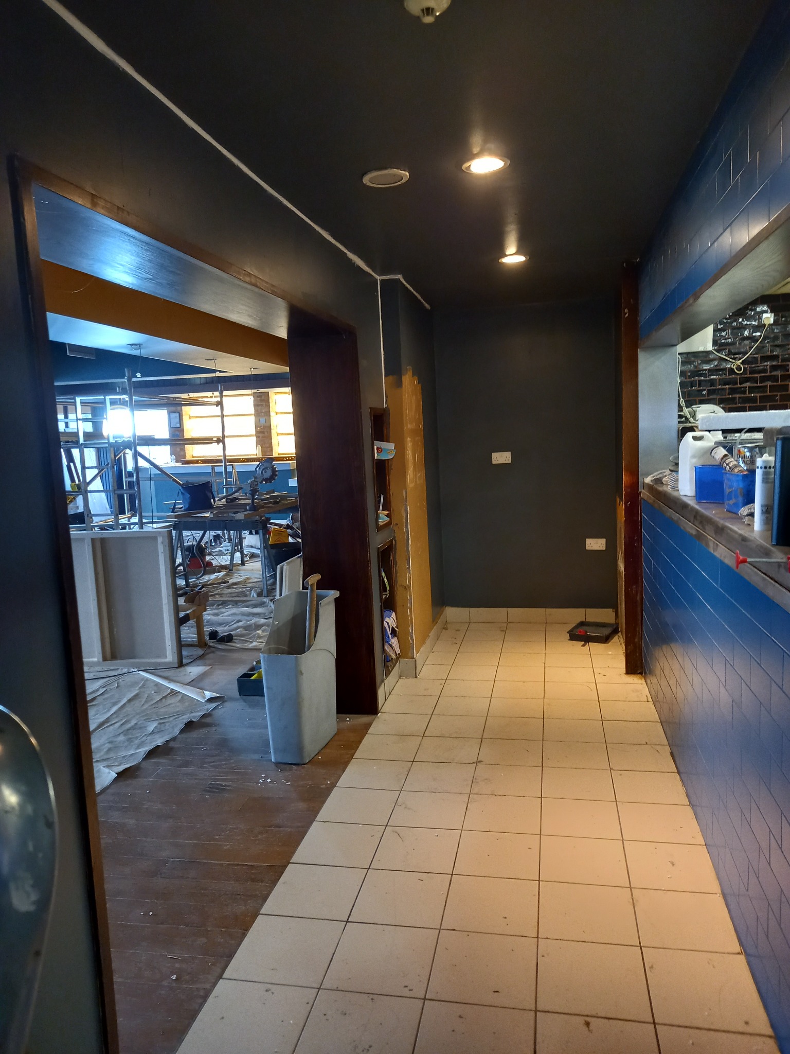 Korytarz w trakcie prac wykończeniowych, widoczne narzędzia, drabina i zabezpieczenia podłogi, kontrastujące z pomalowaną na ciemno ścianą i sufitem oraz wyłożoną kafelkami ścianą po prawej stronie.