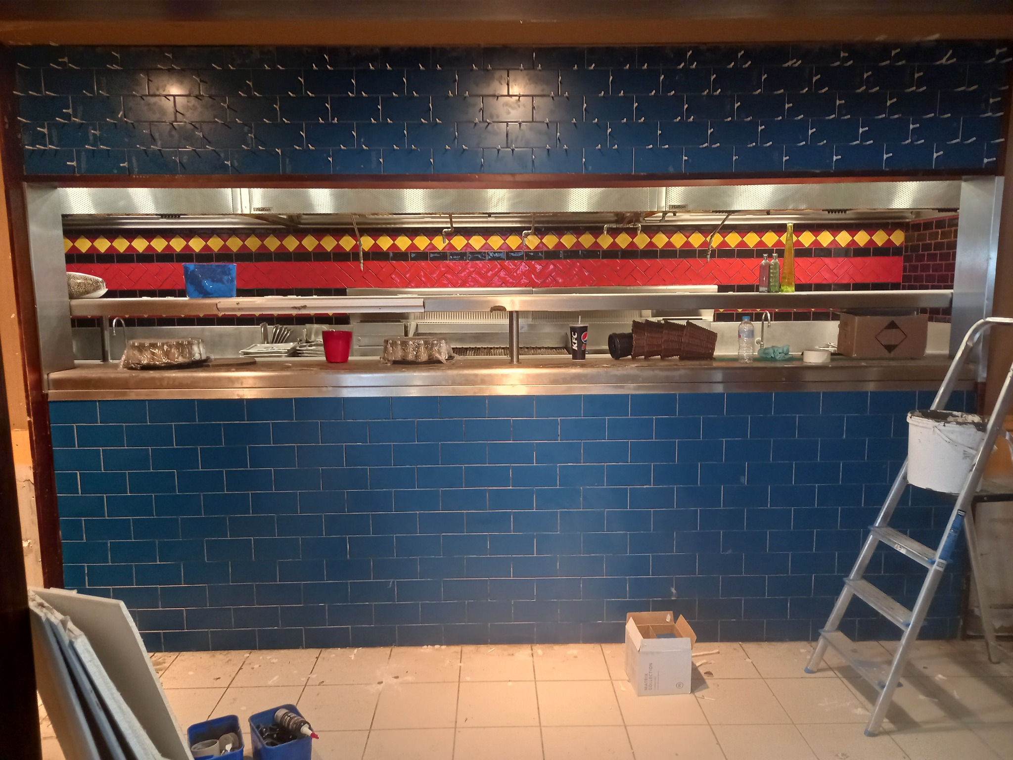 Niebieska ściana z płytek w trakcie układania, widoczne narzędzia i drabina, fragment wnętrza komercyjnego z elementami wykończeniowymi w stylu retro.