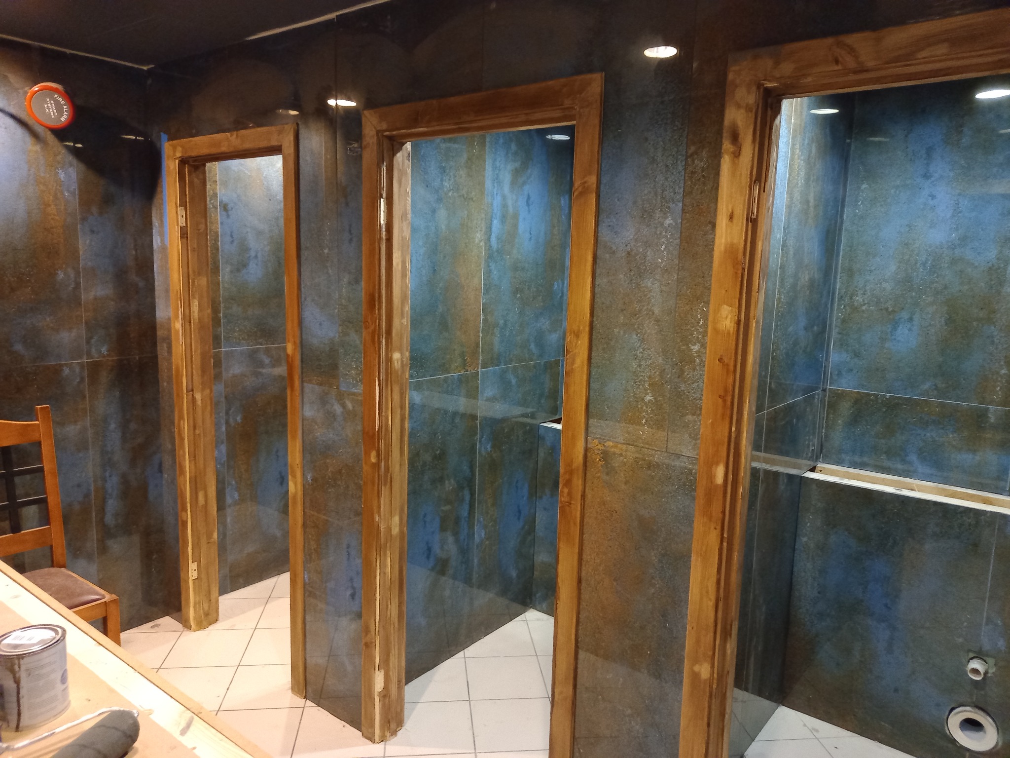 Wnętrze łazienki w trakcie prac wykończeniowych, widoczne trzy drewniane ościeżnice, niebiesko-brązowe płytki na ścianach, biała podłoga z płytek, krzesło i puszka z farbą na pierwszym planie.