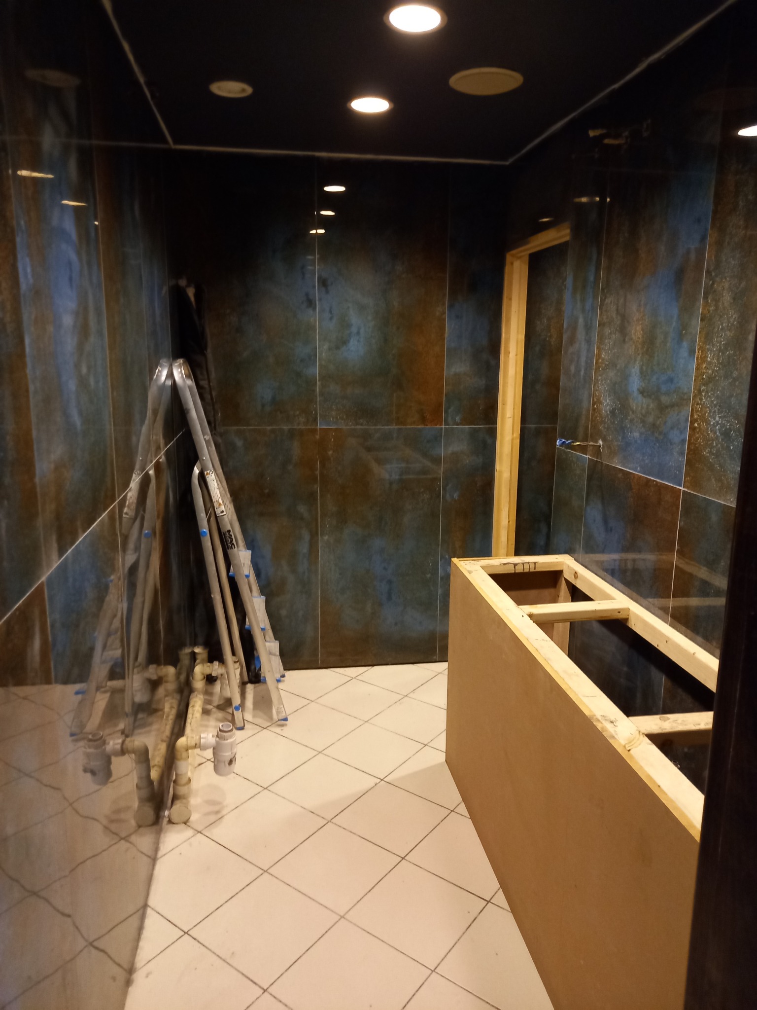 Łazienka w trakcie prac: widoczne płytki imitujące kamień, drabina, rury oraz drewniana konstrukcja pod zabudowę wanny lub prysznica.