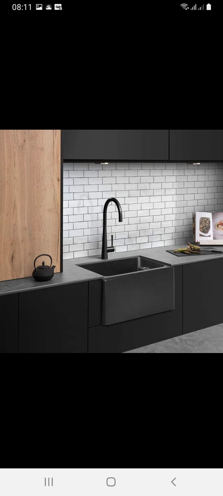 Czarna, ceramiczna umywalka farmerska z baterią w nowoczesnej, minimalistycznej kuchni z białymi, ceglanymi płytkami na ścianie i drewnianą szafką. Dodatkowo czajnik z żeliwa i szparagi na blacie.