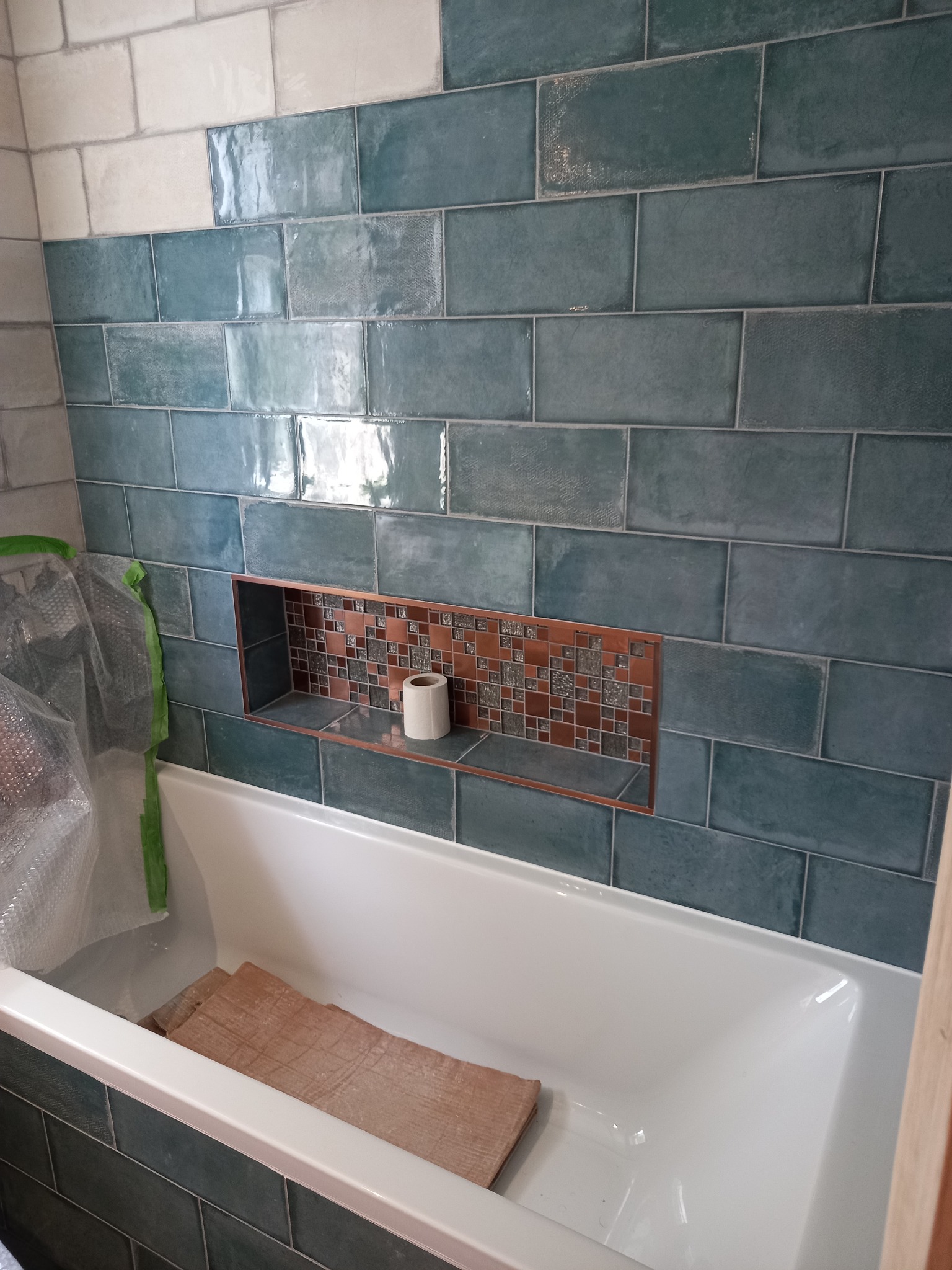 Łazienka w trakcie remontu: ściana wyłożona błyszczącymi, turkusowymi płytkami w stylu cegiełek, wbudowana wnęka z mozaiką w odcieniach brązu, zabezpieczona folią wanna z drewnianymi elementami...