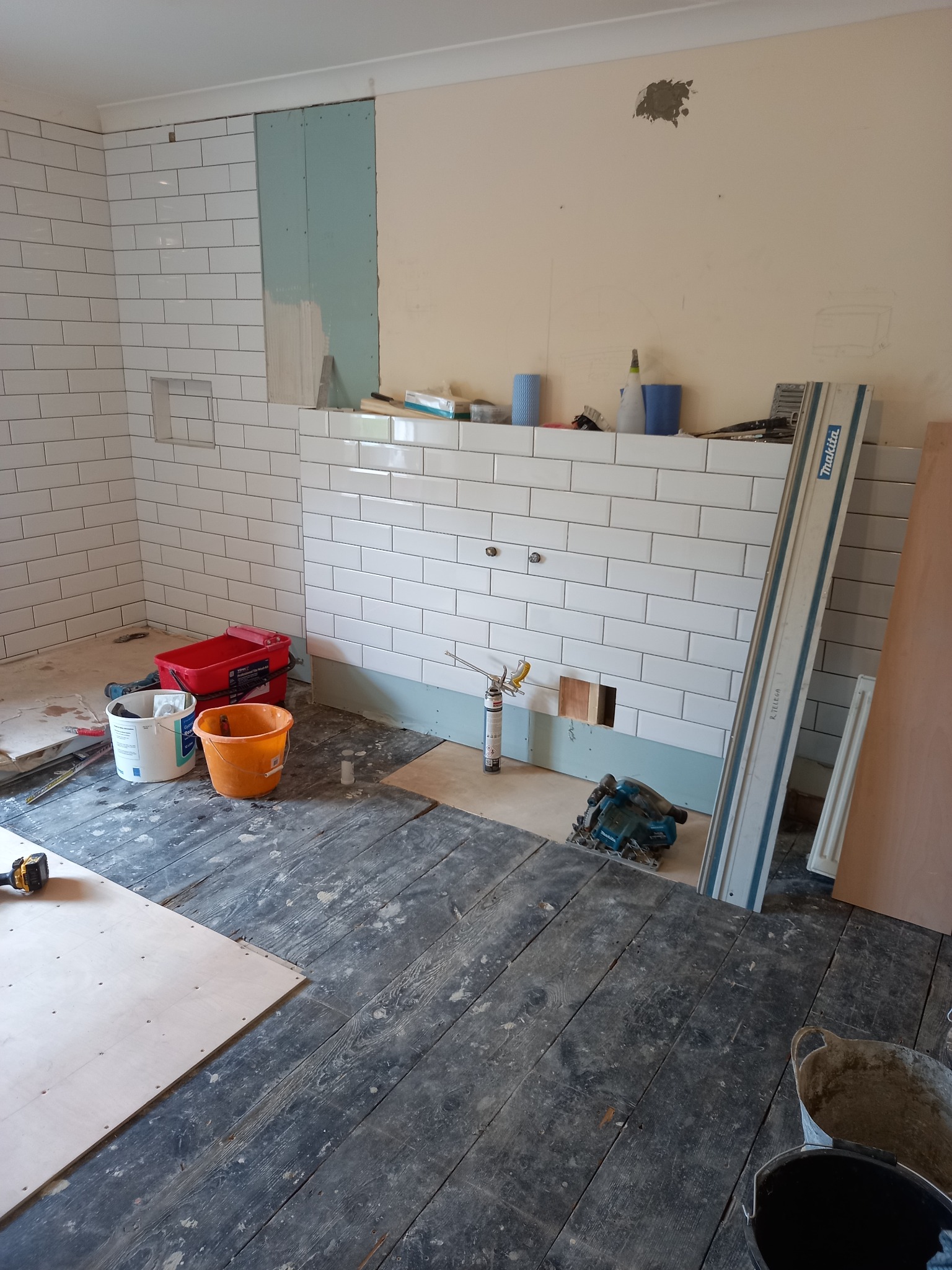 Układanie białych, podłużnych płytek na ścianie łazienki, widoczne narzędzia, wiadro i fragmenty niezakończonej ściany z płytą gipsową.