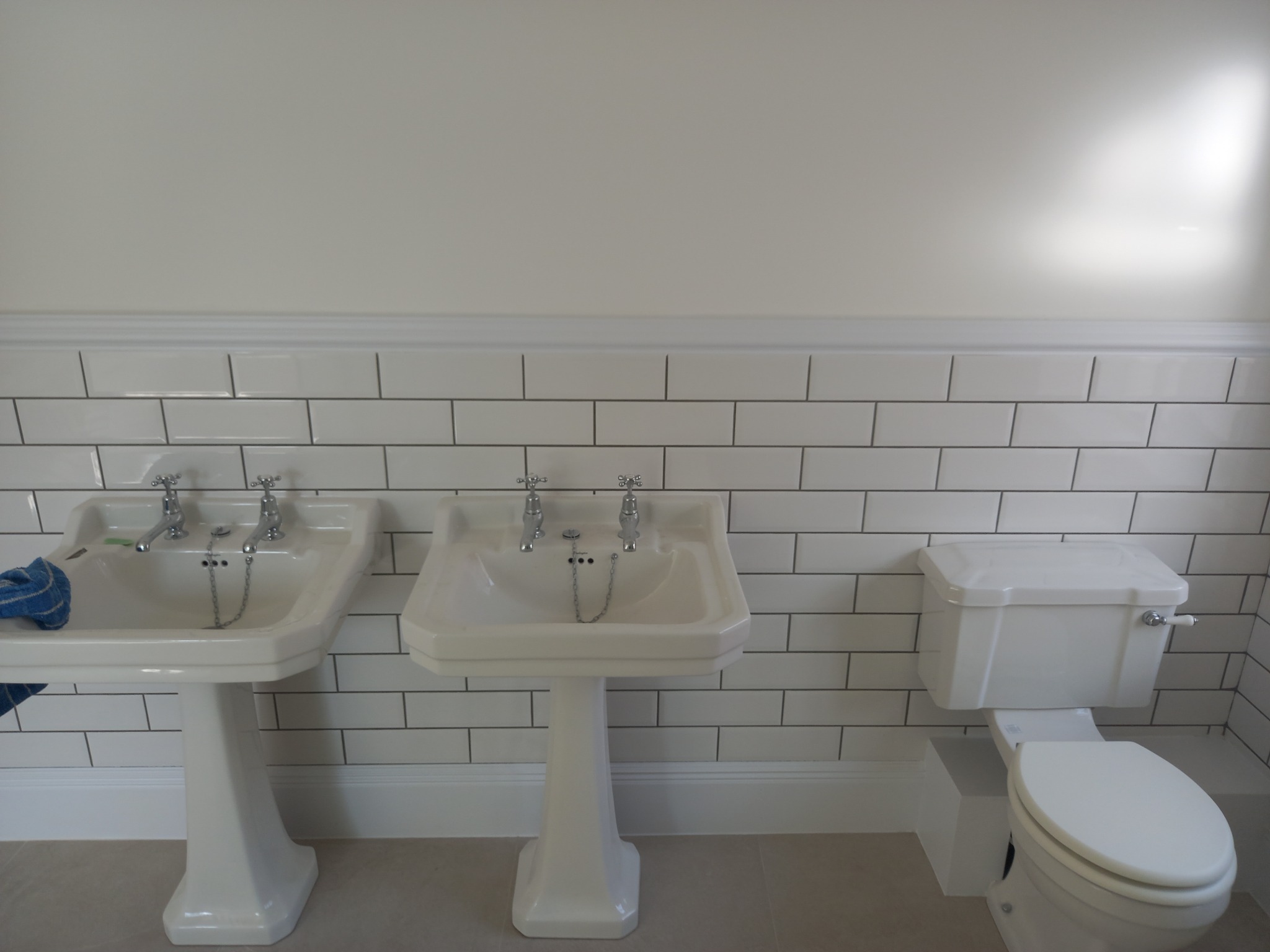 Dwie białe umywalki na postumentach i toaleta w wykończonej łazience z białymi płytkami metra i jasnymi ścianami.