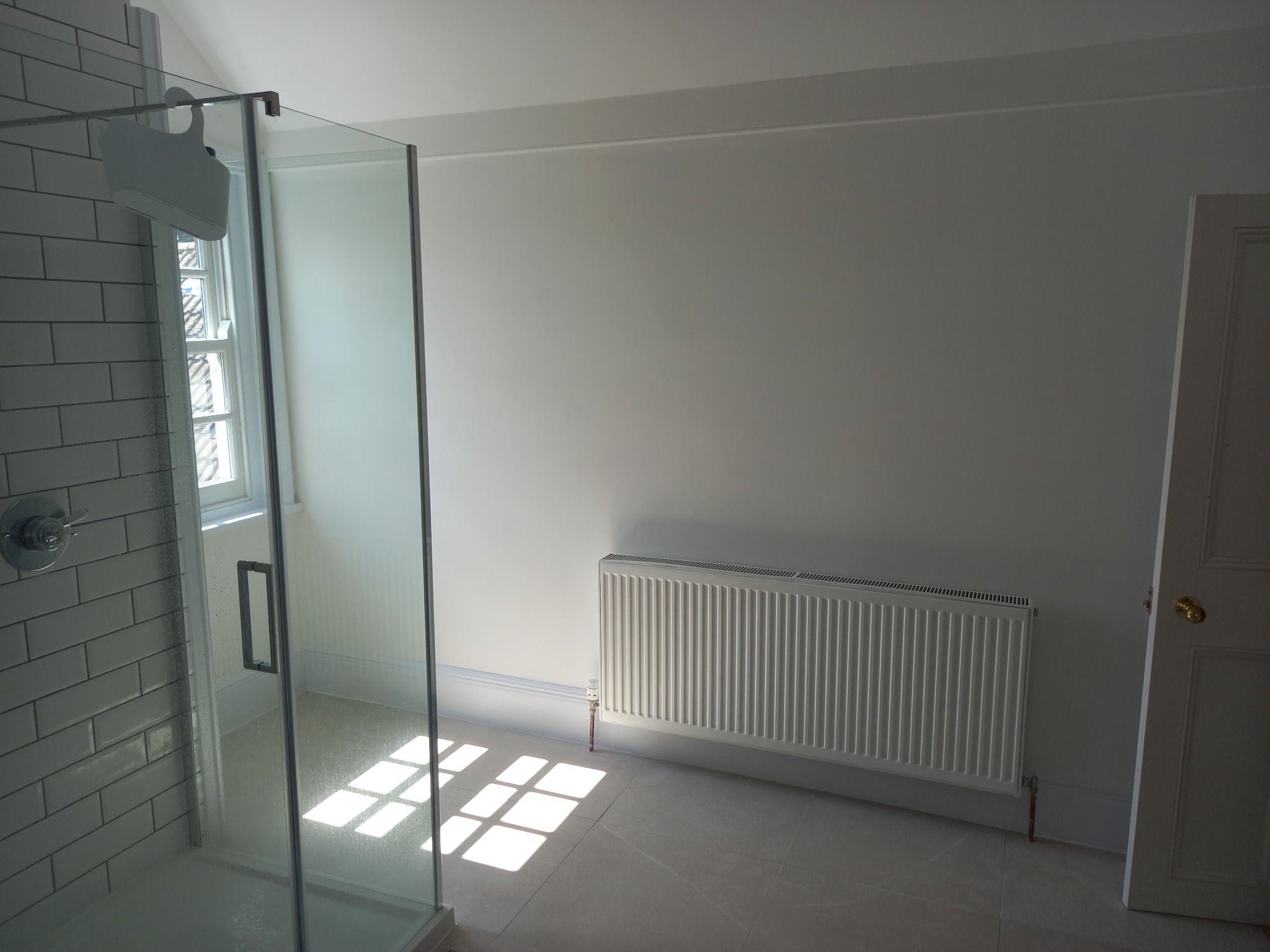 Jasna łazienka po renowacji z kabiną prysznicową ze szklaną ścianką, grzejnikiem panelowym i promieniami słońca na podłodze.