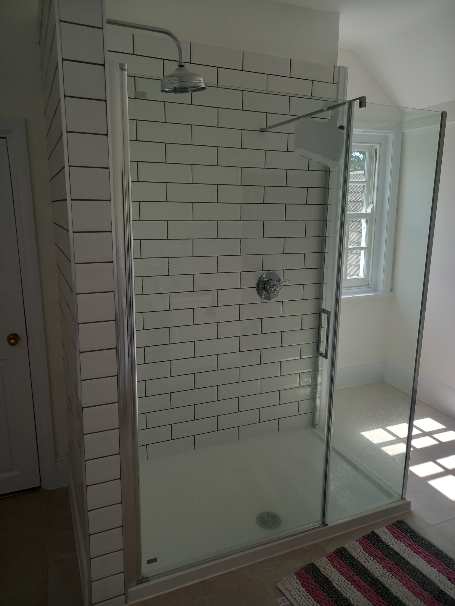 Nowoczesna kabina prysznicowa z białymi, prostokątnymi płytkami ułożonymi w cegiełkę, szklanymi drzwiami i chromowaną armaturą, widok na okno z parapetem.