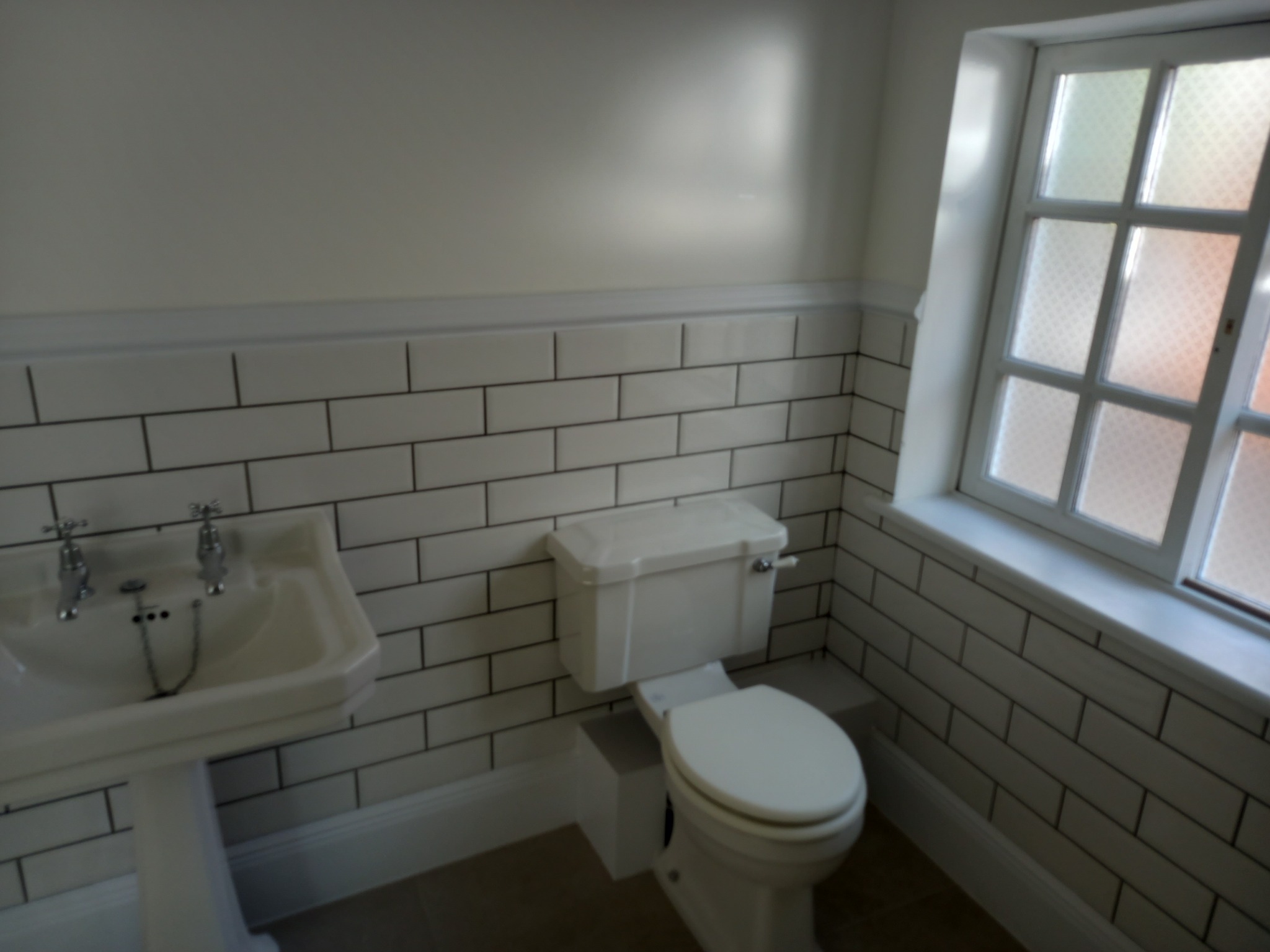 Jasna łazienka z białą umywalką na postumencie, toaletą i oknem z mleczną szybą, wykończona białymi płytkami typu metro z ciemną fugą.