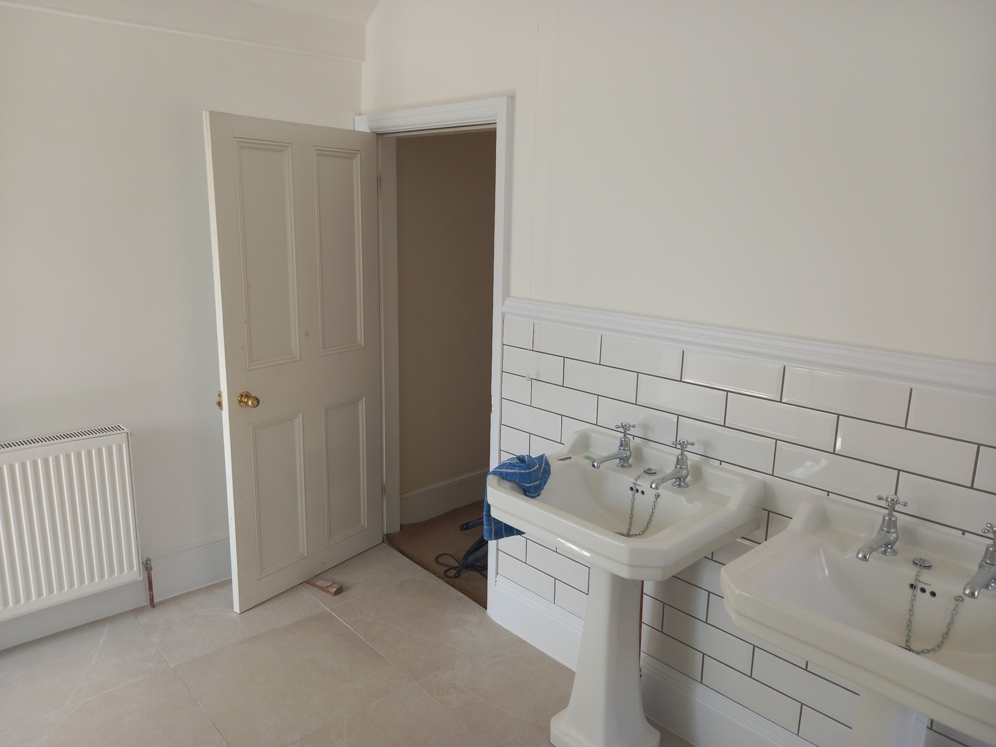 Jasna łazienka z dwoma umywalkami na postumentach, białe płytki na ścianach i otwarta biała drewniana rama drzwiowa, widoczny grzejnik.