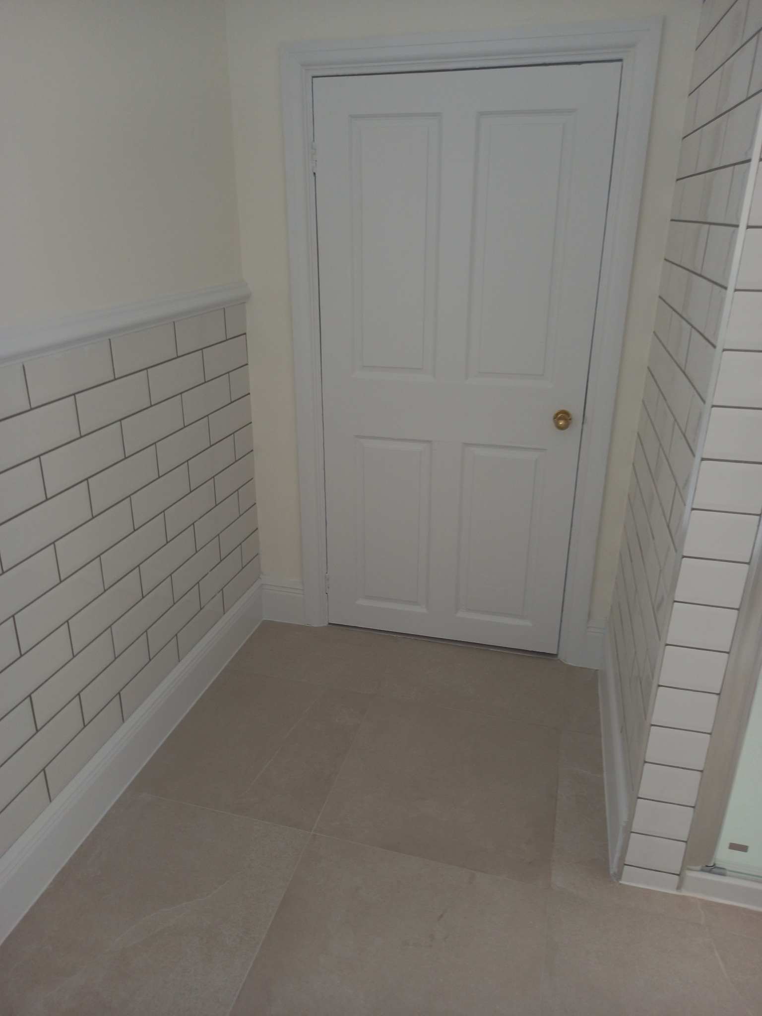 Wykończona łazienka z białymi, prostokątnymi płytkami ułożonymi w cegiełkę na ścianach do połowy wysokości, szarą podłogą z płytek oraz białymi drzwiami z mosiężną klamką.
