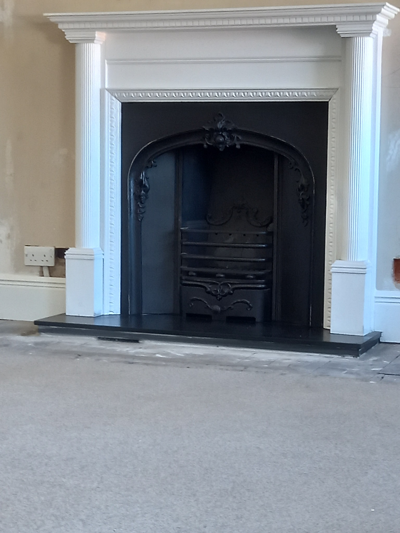 Klasyczny, czarny kominek z ozdobnymi detalami, otoczony białym, kolumnowym portalem w trakcie prac wykończeniowych. Widoczne gniazdko elektryczne na ścianie. Podłoga częściowo zakryta. Ściana...