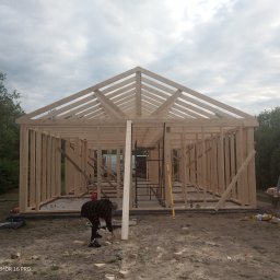 Marcin Zagłoba Usługi Budowlane - Wznoszenie drewnianej konstrukcji domu szkieletowego z widocznym szkieletem dachu, pracownik przy ziemi oraz drugi z taczką w tle.