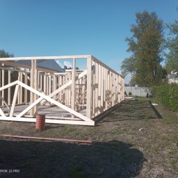 Marcin Zagłoba Usługi Budowlane - Drewniana konstrukcja szkieletowa domu w trakcie budowy, widoczny szkielet ścian i dachu na tle błękitnego nieba, z widocznym fragmentem samochodu dostawczego po lewej stronie.