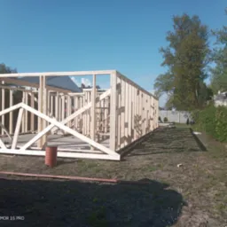 Drewniana konstrukcja szkieletowa domu w trakcie budowy, widoczny szkielet ścian i dachu na tle błękitnego nieba, z widocznym fragmentem samochodu dostawczego po lewej stronie.