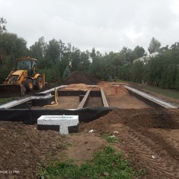 Marcin Zagłoba Usługi Budowlane - Przygotowanie terenu pod budowę: wykopane fundamenty z izolacją, widoczny żółty traktor i hałdy ziemi na tle zieleni i pochmurnego nieba.