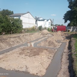 Marcin Zagłoba Usługi Budowlane - Zalane betonem wykopy pod fundamenty domu jednorodzinnego, widoczny fragment zbrojenia, w tle budynki mieszkalne i czerwony samochód dostawczy.