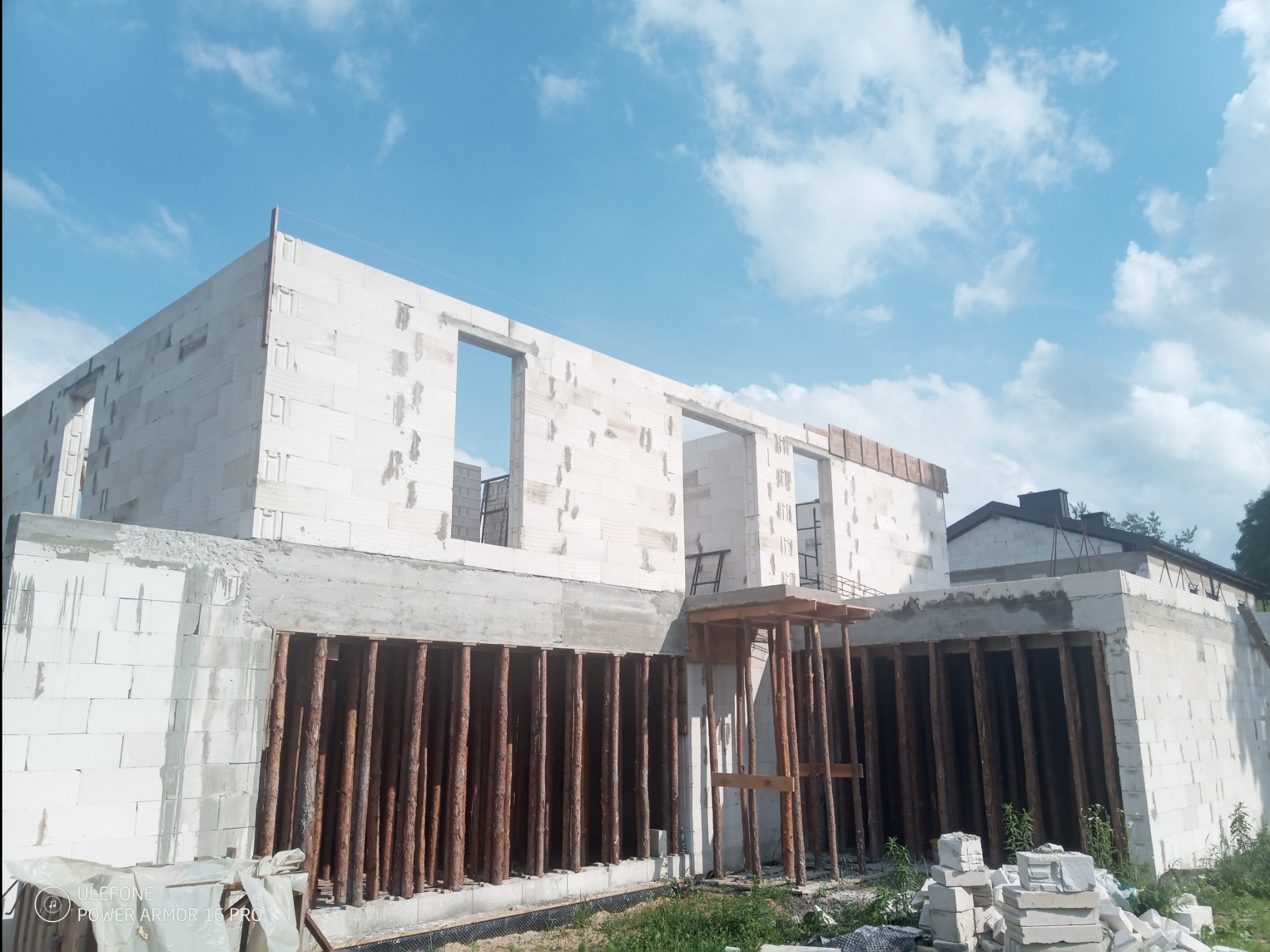 Wznoszenie ścian domu z białych bloczków, widoczne otwory okienne i drewniane stemple podtrzymujące konstrukcję fundamentów, błękitne niebo w tle.