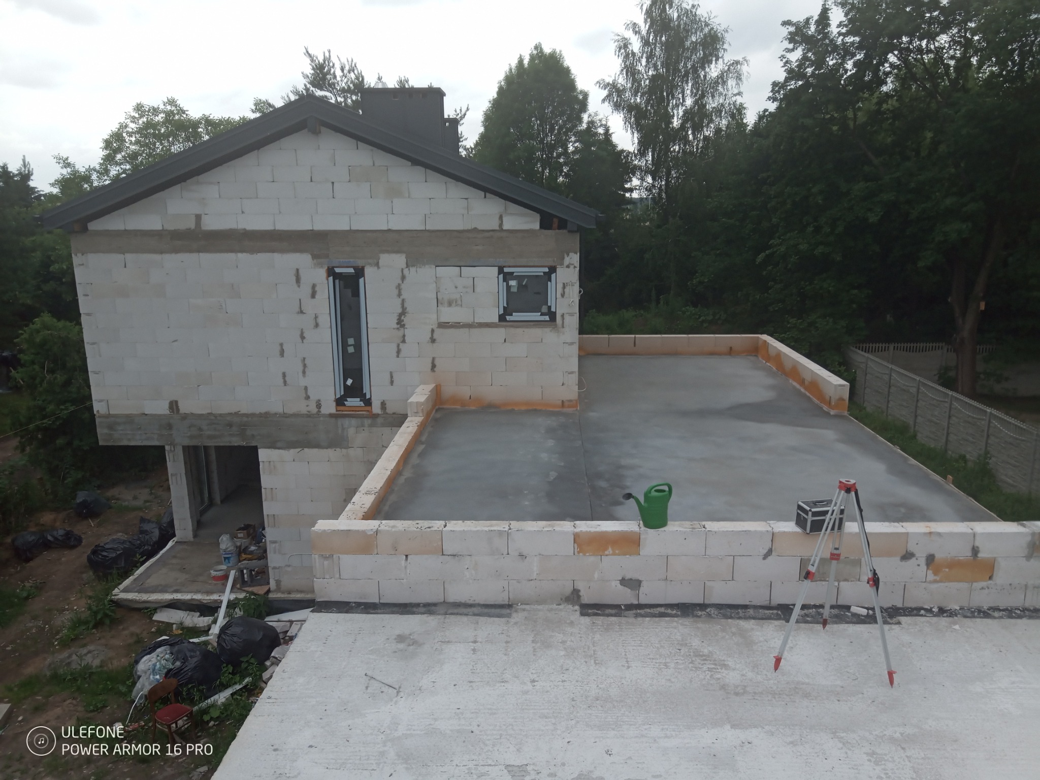 Budowa domu z białych bloczków: surowy stan, widoczne okna, świeżo wylany strop z zieloną konewką i niwelatorem na nim, otoczenie drzew