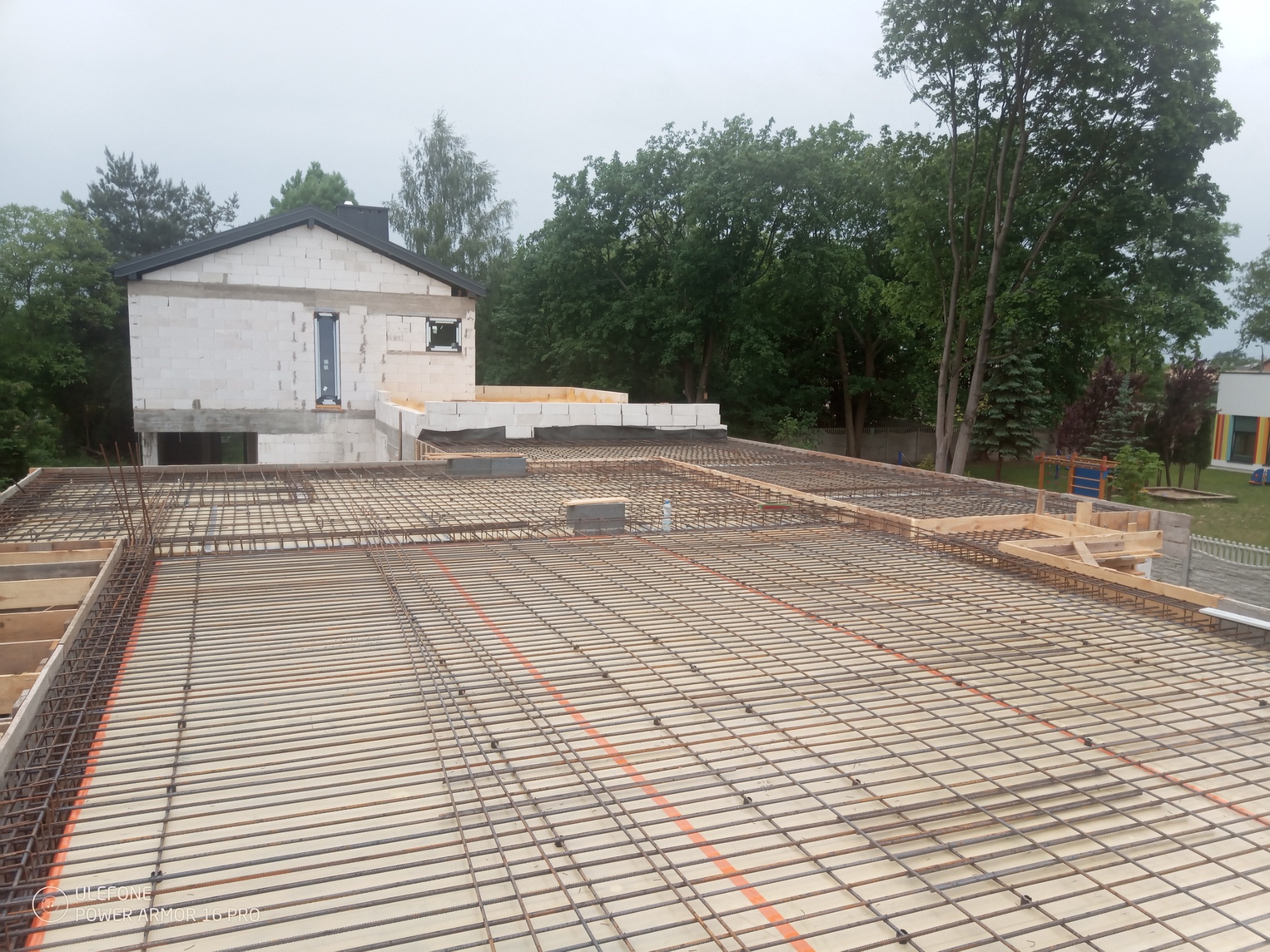 Zbrojenie stropu budynku w trakcie budowy, widok z góry na siatkę z prętów stalowych, w tle ściany z białego bloczka i zielone drzewa.