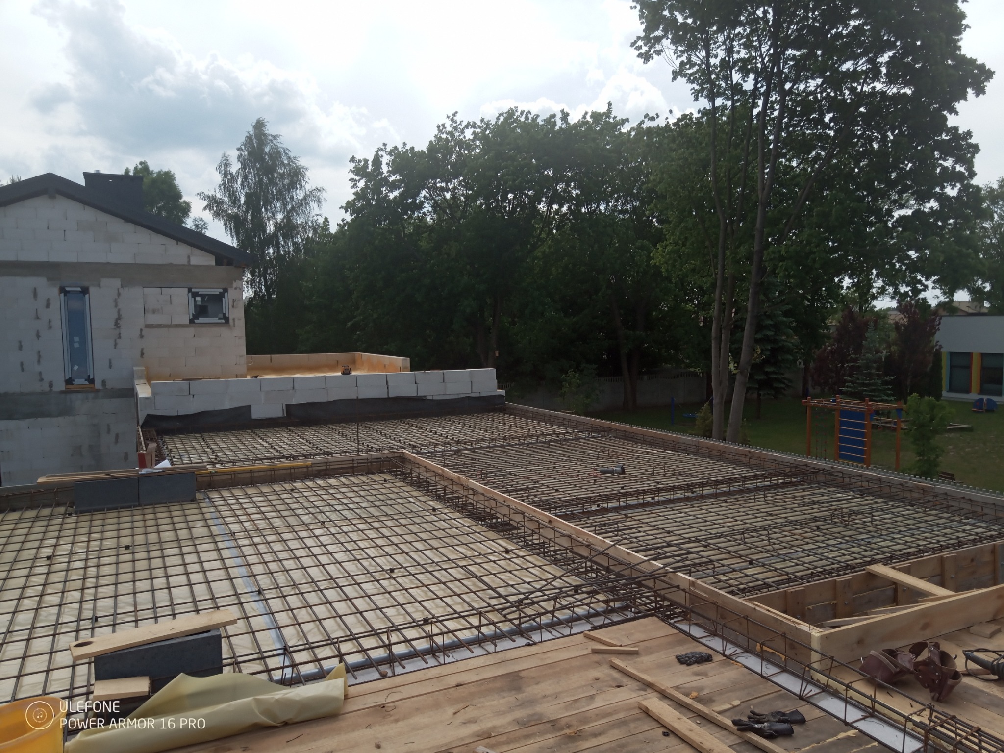 Zbrojenie stropu budynku w trakcie budowy, widoczne drewniane deskowanie, siatka z prętów stalowych i fragment muru z pustaków.