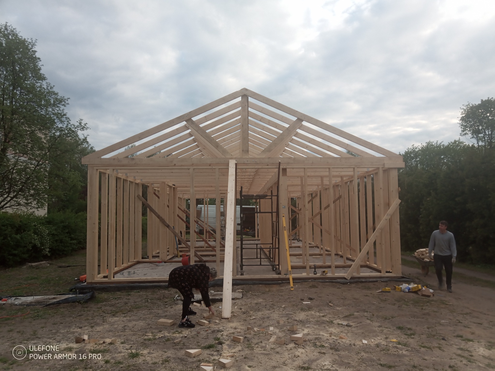 Wznoszenie drewnianej konstrukcji domu szkieletowego z widocznym szkieletem dachu, pracownik przy ziemi oraz drugi z taczką w tle.