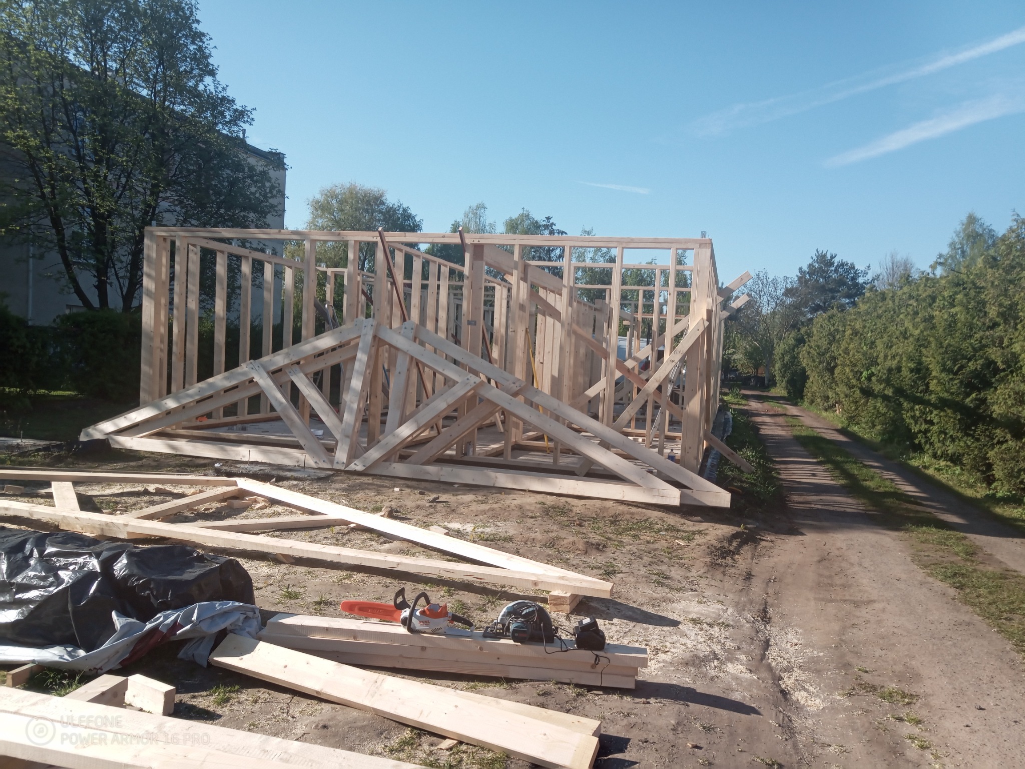 Konstrukcja szkieletowa drewnianego domu w trakcie budowy, widoczne elementy ścian i więźby dachowej, piła łańcuchowa i elektronarzędzia leżą na deskach, w tle droga gruntowa i budynek mieszkalny.